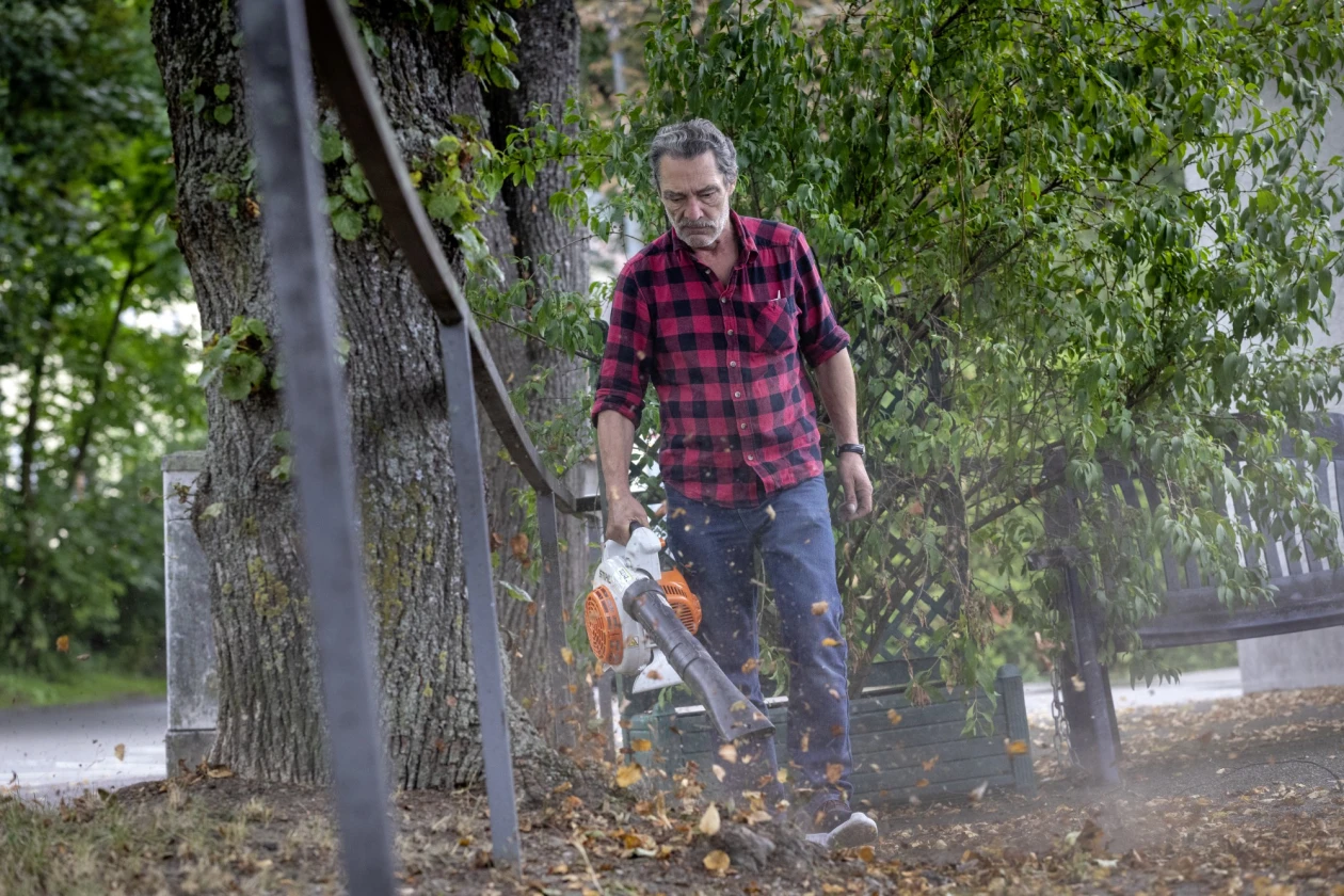 Ein Mann in kariertem Hemd arbeitet mit einem Laubsauger, umgeben von B&auml;umen und herbstlichen Bl&auml;ttern.