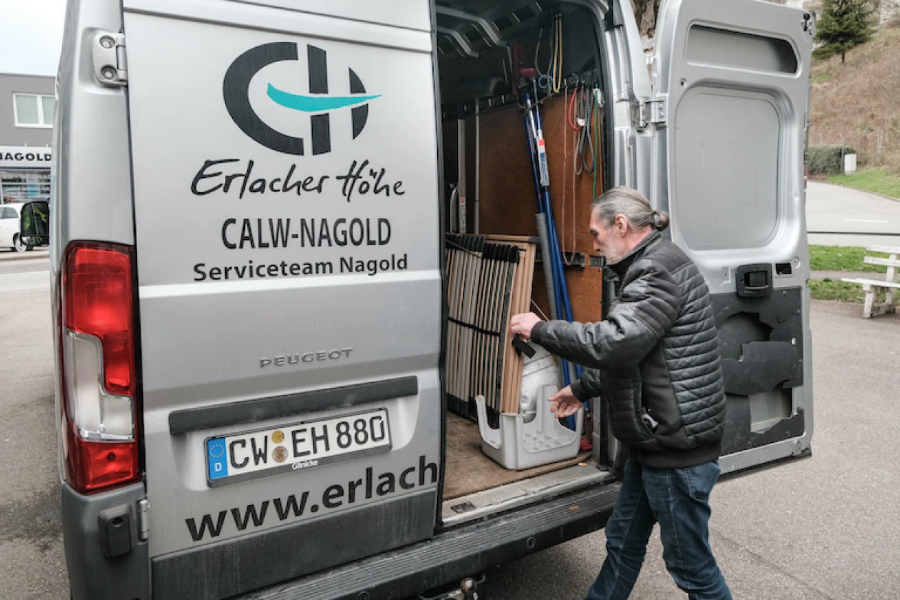 Ein Mann in einer Daunenjacke steht vor der Ladefl&auml;che eines silbernen Transporters des Serviceteams Nagold von der Erlacher H&ouml;he. Auf der Ladefl&auml;che ist ein Holzbettgestell und weitere Materialien zu sehen.