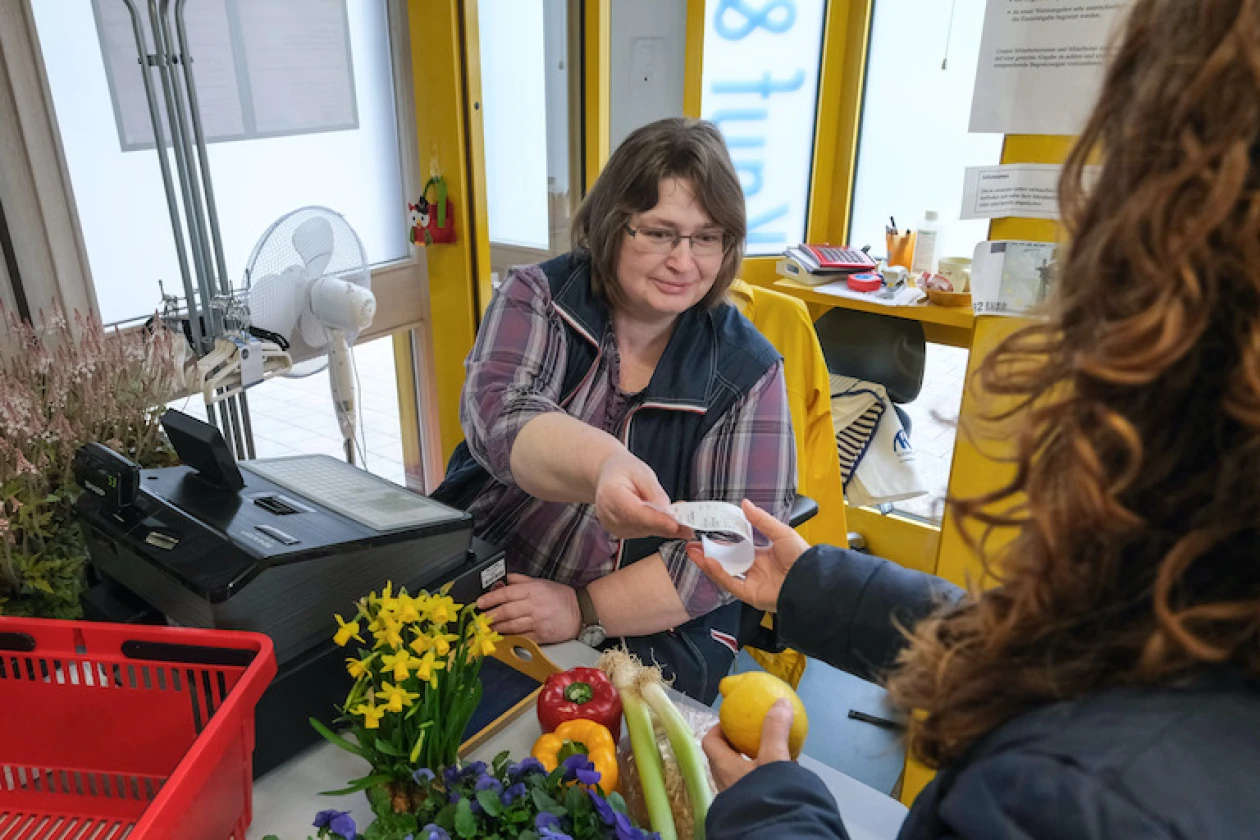 Eine Frau in kariertem Hemd und Weste arbeitet an der Kasse und reicht einer Kundin, die mit dem R&uuml;cken zur Kamera steht, eine Quittung. Auf dem Tresen liegen Blumen und Obst.