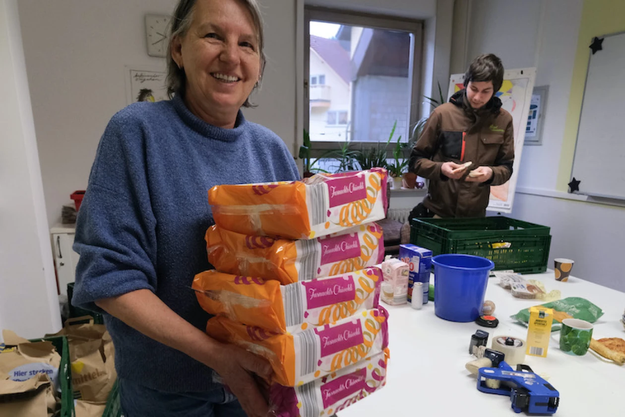 Eine l&auml;chelnde Frau in einem blauen Pullover h&auml;lt einen Stapel orangefarbener Verpackungen in der Hand, w&auml;hrend im Hintergrund eine weitere Person arbeitet. Auf dem Tisch sind Lebensmittel zu sehen, die ausgezeichnet werden.