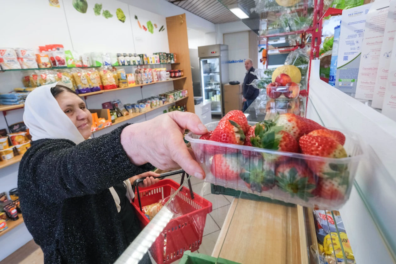 Eine Frau mit wei&szlig;em Kopftuch w&auml;hlt Erdbeeren aus einer Obsttheke in einem Laden aus und h&auml;lt einen roten Einkaufskorb in der Hand. Im Hintergrund sind weitere Lebensmittel zu sehen.