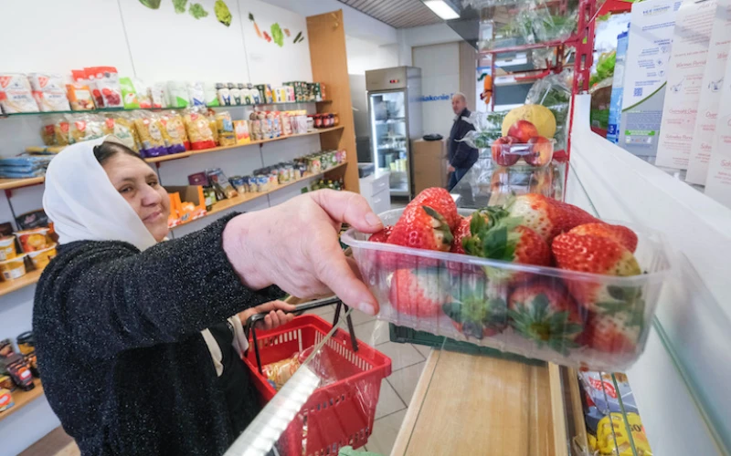 Eine Frau mit wei&szlig;em Kopftuch w&auml;hlt Erdbeeren aus einer Obsttheke in einem Laden aus und h&auml;lt einen roten Einkaufskorb in der Hand. Im Hintergrund sind weitere Lebensmittel zu sehen.
