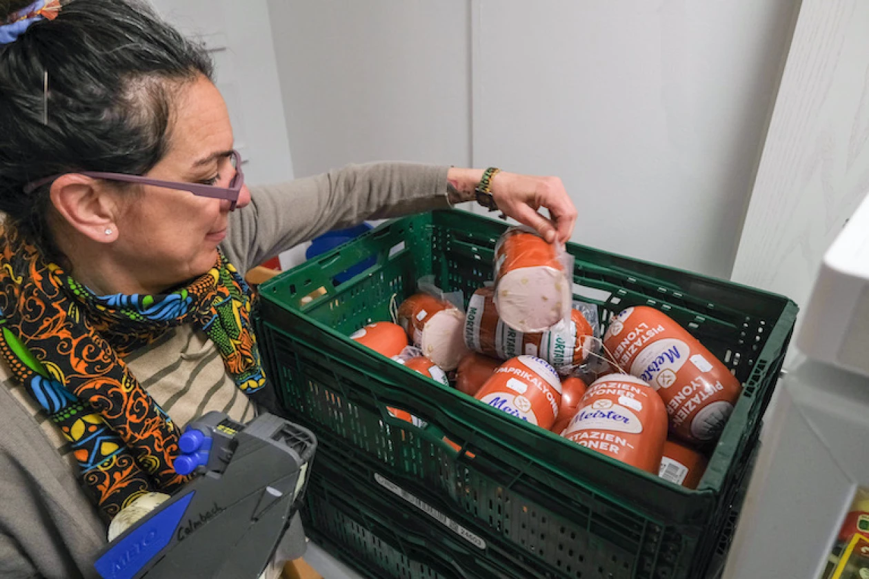 Eine Frau mit Brille und buntem Schal nimmt eine in Folie verpackte Wurst aus einer gr&uuml;nen Kiste, in der sich noch weitere Wurst befindet, und zeichnet diese mit dem Preis aus.