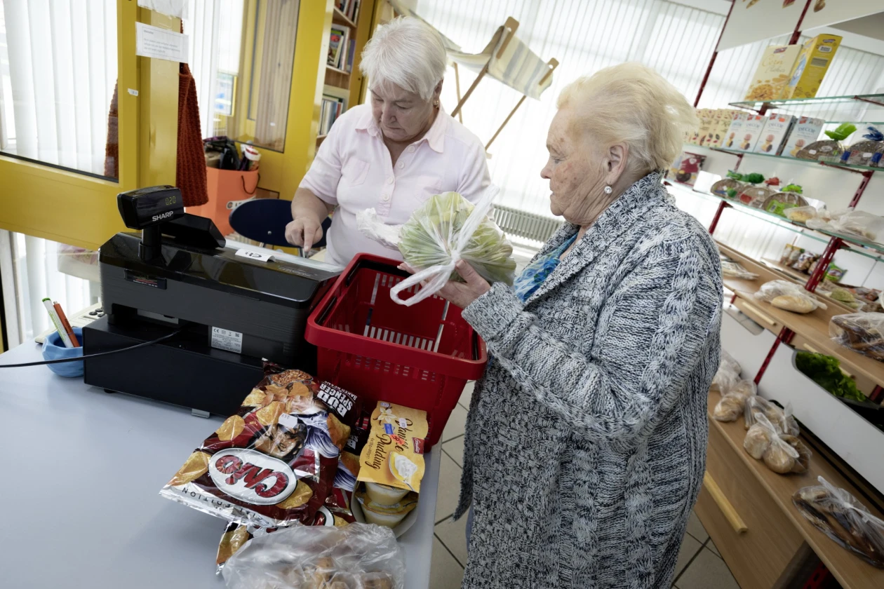 Eine Frau in wei&szlig;er Bluse steht an der Kasse, w&auml;hrend eine andere Frau im grauen Pullover mit einem roten Einkaufskorb neben ihr wartet. Im Hintergrund sind Regale mit Backwaren zu sehen.