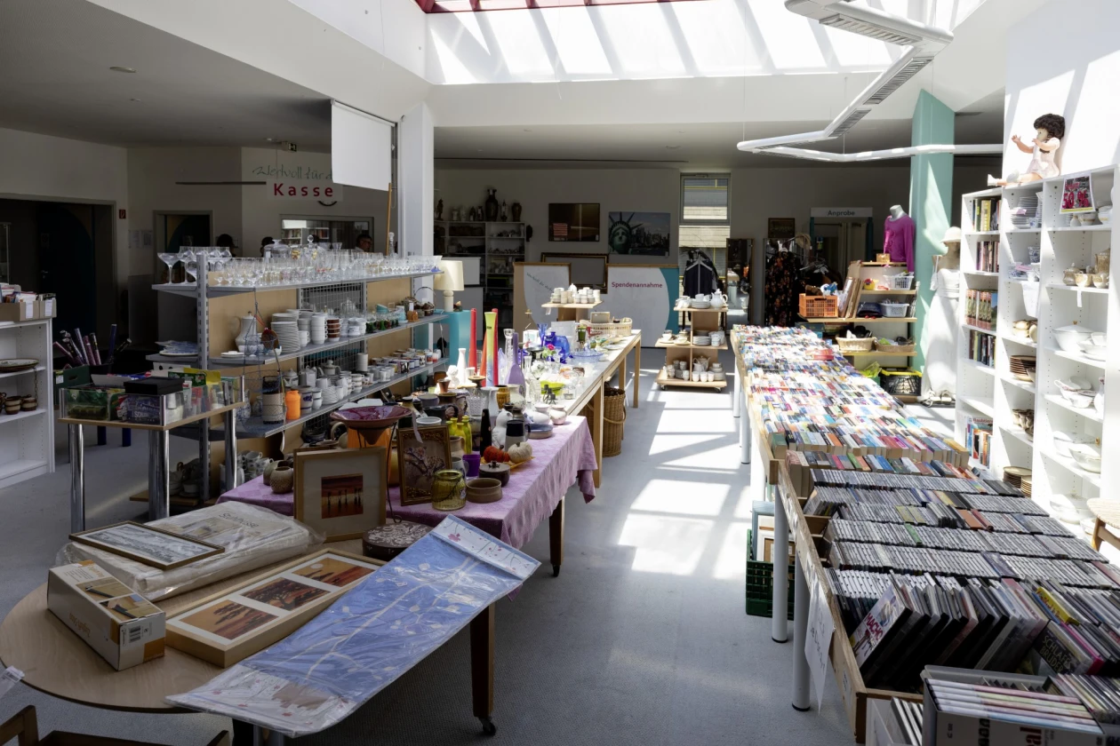 Gro&szlig;er Verkaufsraum mit mehreren Tischen und Regalen, die mit verschiedenen Waren best&uuml;ckt sind, darunter B&uuml;cher, Gl&auml;ser, Geschirr und andere Haushaltsgegenst&auml;nde.