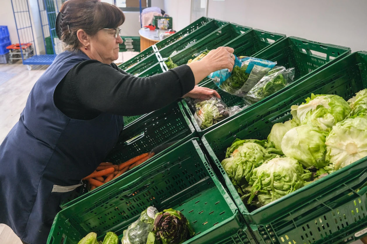 Zu sehen ist eine Frau mit Brille, umgeben von zahlreichen gr&uuml;nen Kisten, die mit verschiedenen Gem&uuml;sesorten gef&uuml;llt sind.