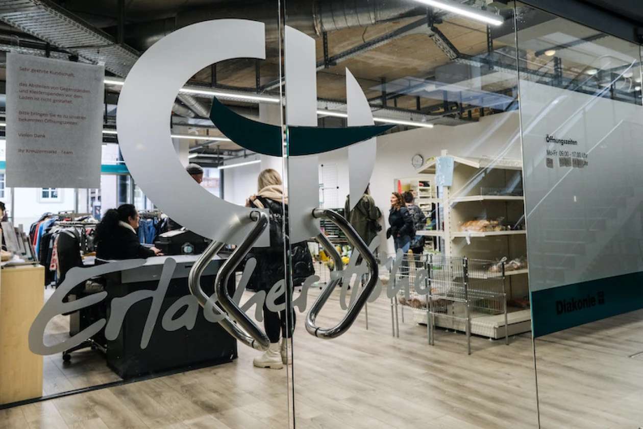 Zu sehen ist eine gro&szlig;e Glasfront mit Eingangst&uuml;re in ein Ladengesch&auml;ft mit gro&szlig;em Schriftzug und dem Logo der &bdquo;Erlacher H&ouml;he&ldquo;. Durch die Glasfront sind Lebensmittelregale und Kleiderst&auml;nder sowie mehrere Personen zu sehen.