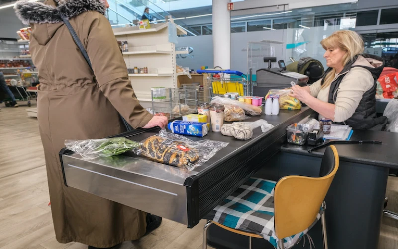 Eine Frau an einer Kasse kassiert den Einkauf einer Kundin ab. Verschiedene Lebensmittel liegen auf dem Kassenband. Eine gelber Stuhl ist im Vordergrund sichtbar.
