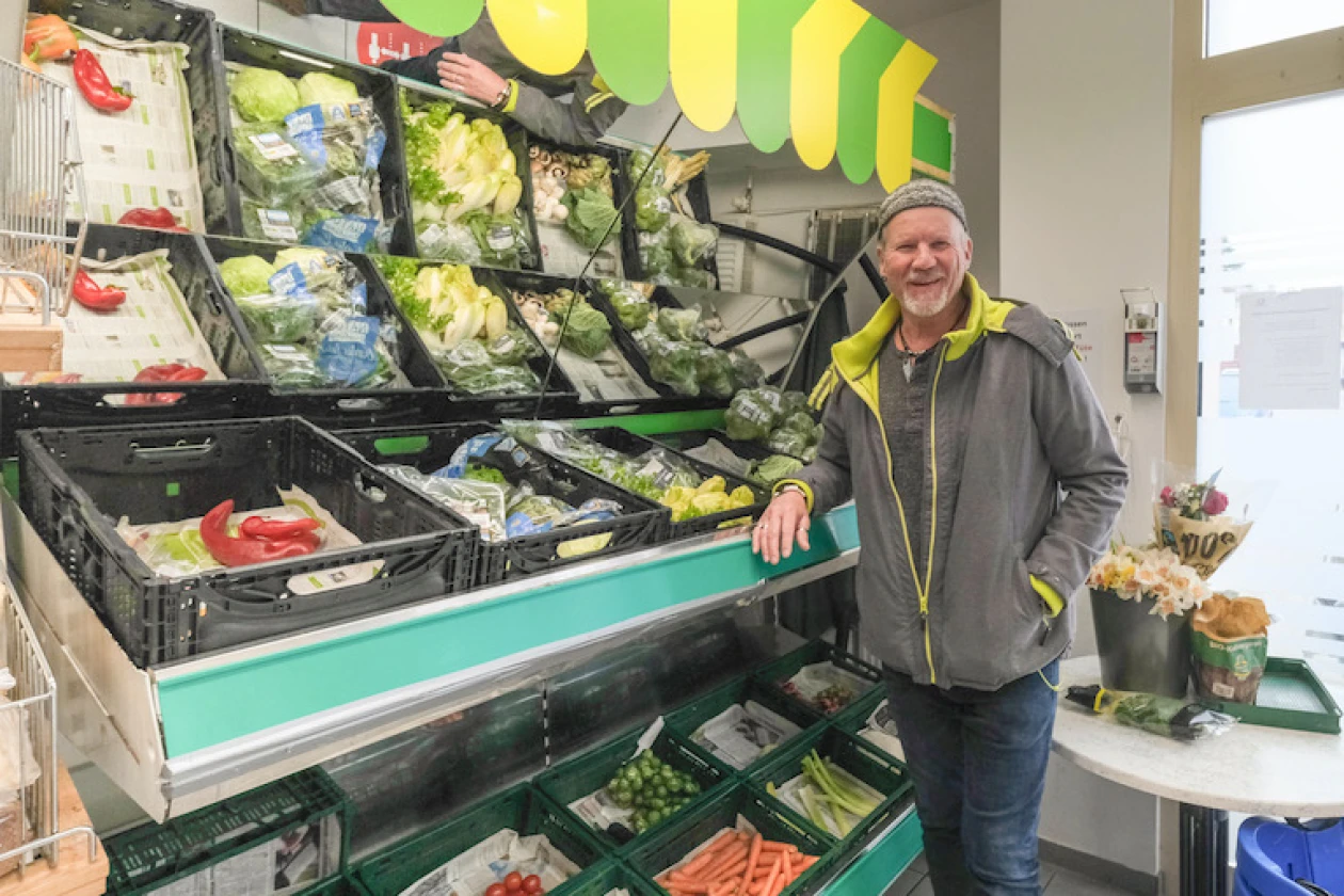 Ein Mann in grauer Jacke und M&uuml;tze lehnt sich an ein Gem&uuml;seregal. Das Gem&uuml;se ist in verschiedenen Kisten sortiert und pr&auml;sentiert eine bunte Vielfalt.