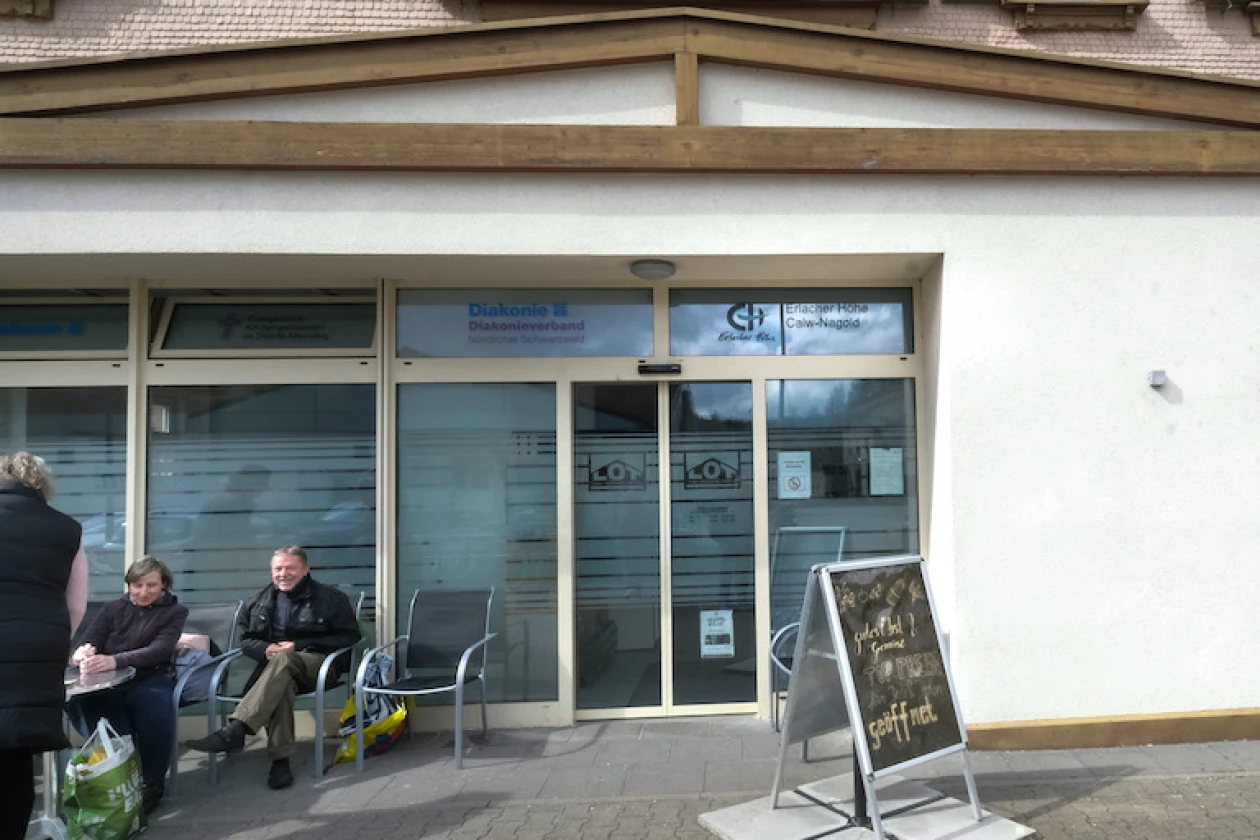 Zwei Personen sitzen auf St&uuml;hlen vor einem Geb&auml;ude mit moderner Glasfront und Holzverkleidung. Rechts daneben steht eine Infotafel. Auf der Glasfront ist der Schriftzug &bdquo;Erlacher H&ouml;he Calw-Nagold&ldquo; zu lesen.