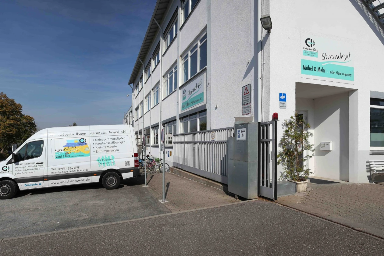 Zu sehen ist das ein wei&szlig;es Geb&auml;ude mit der Aufschrift "Erlacher H&ouml;he Strandgut". Links vom Geb&auml;ude ist Parkfl&auml;che, auf dem ein wei&szlig;er Transporter steht.