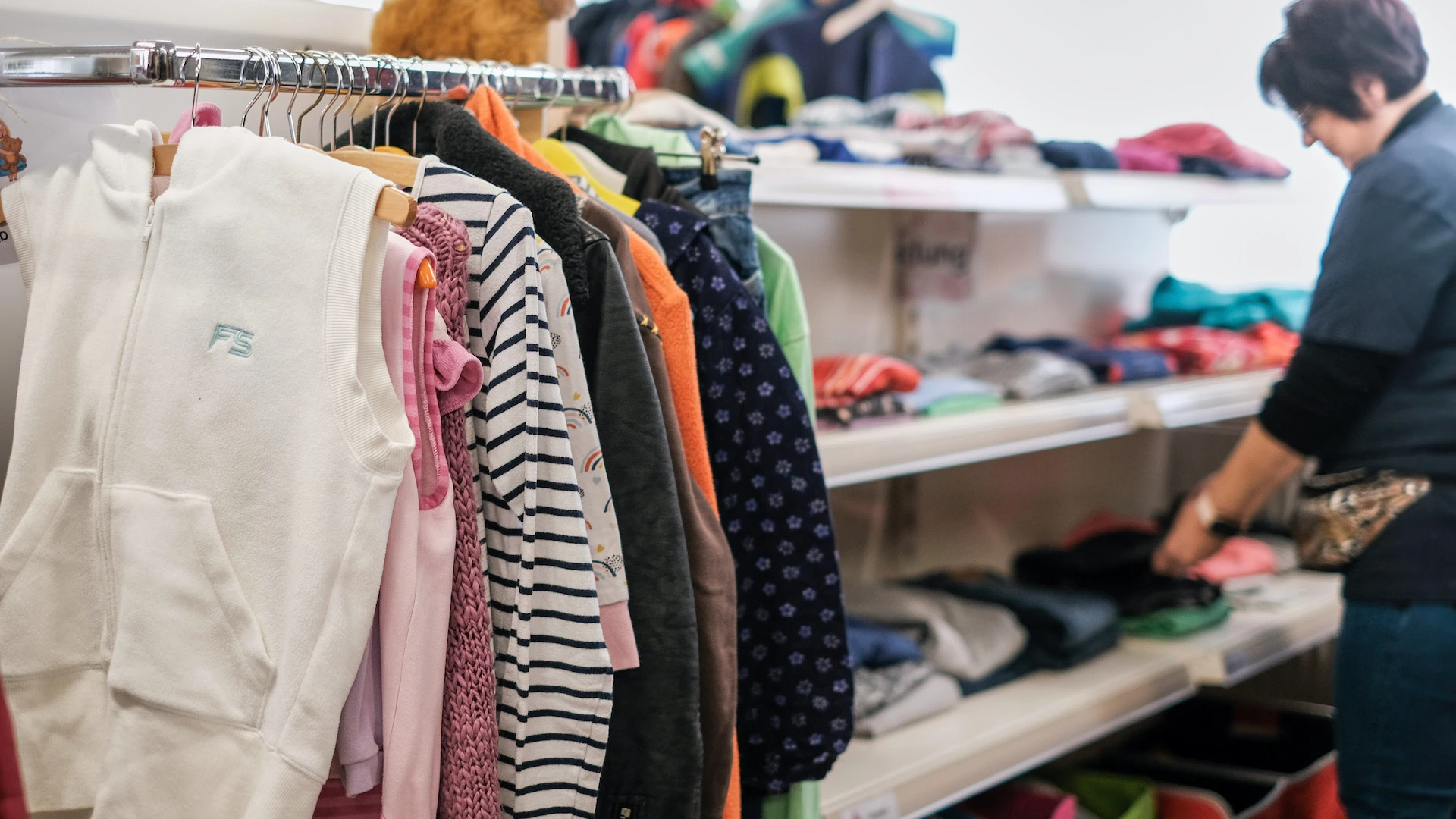Kleiderst&auml;nder mit verschiedenfarbigen Kleidungsst&uuml;cken in einem Ladengesch&auml;ft. Eine Frau steht im Hintergrund und sortiert Textilien in Regalen.