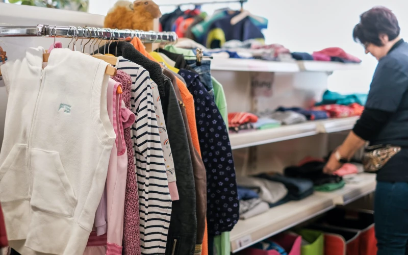 Kleiderst&auml;nder mit verschiedenfarbigen Kleidungsst&uuml;cken in einem Ladengesch&auml;ft. Eine Frau steht im Hintergrund und sortiert Textilien in Regalen.