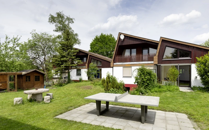 Vor einer Gruppe kleiner Wohnh&auml;user und einem Gartenh&auml;uschen ist eine Rasenfl&auml;che mit einer Tischtennisplatte zu sehen.