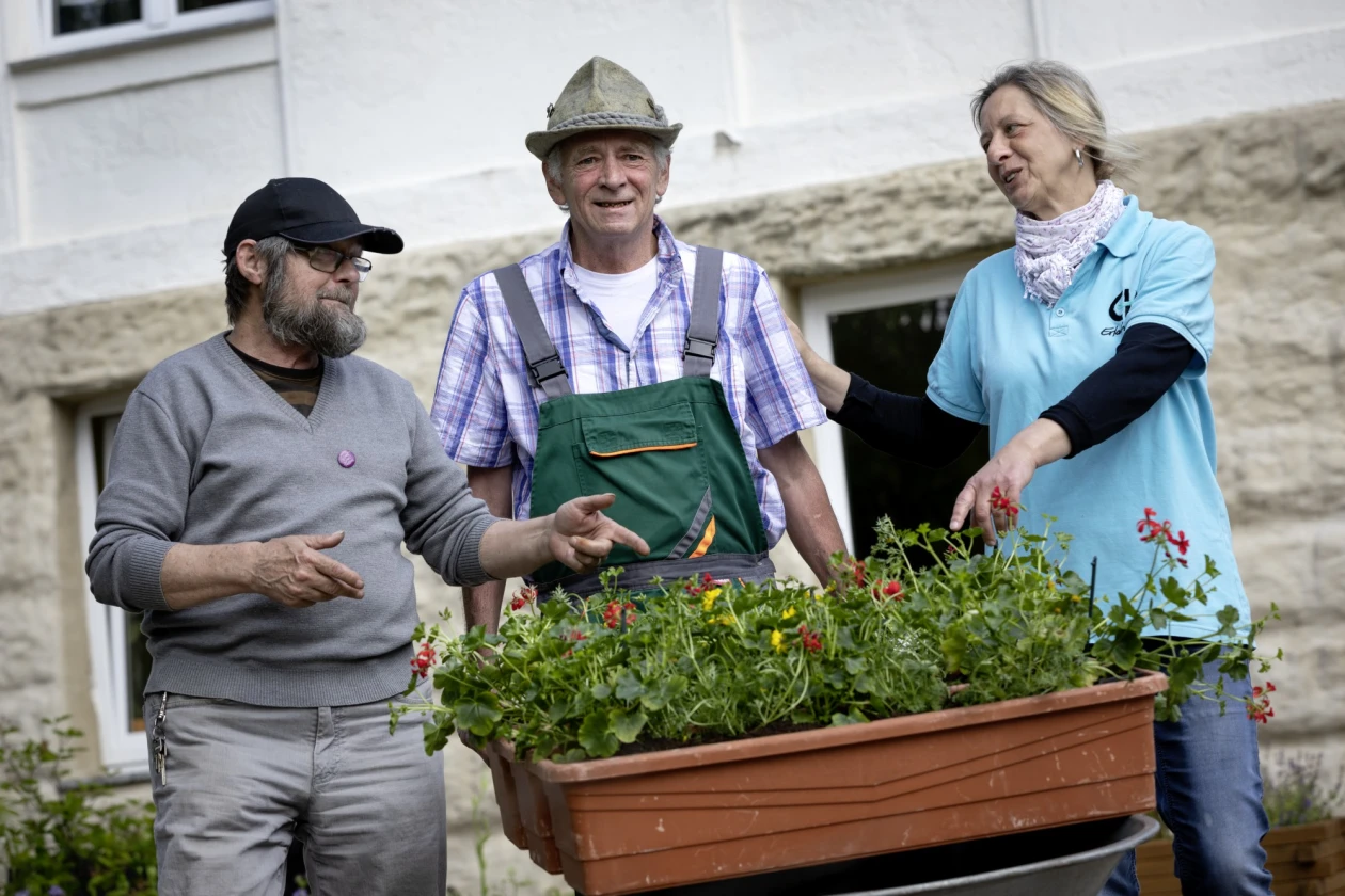 Drei Personen stehen an einem Blumenkasten. Ein Mann in Latzhose und Hut steht in der Mitte, flankiert von einem b&auml;rtigen Mann mit M&uuml;tze links und einer Frau in hellblauem T-Shirt mit Logo der Erlacher H&ouml;he rechts.