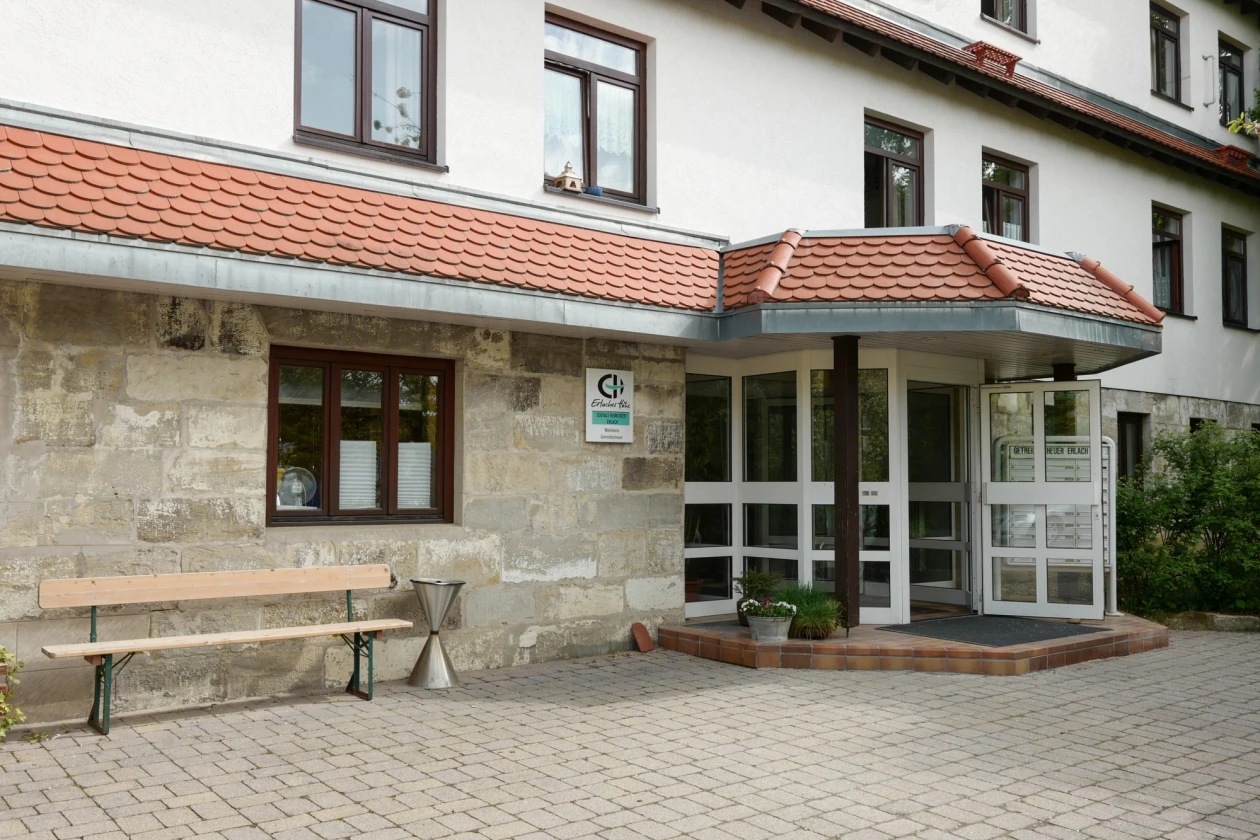Eingangsfassade eines wei&szlig;en Geb&auml;udes mit r&ouml;tlichem Ziegeldach und Klinkersteinfassade, ausgestattet mit einer &uuml;berdachten Glaseingangst&uuml;r, davor eine Sitzbank auf gepflastertem Vorplatz.