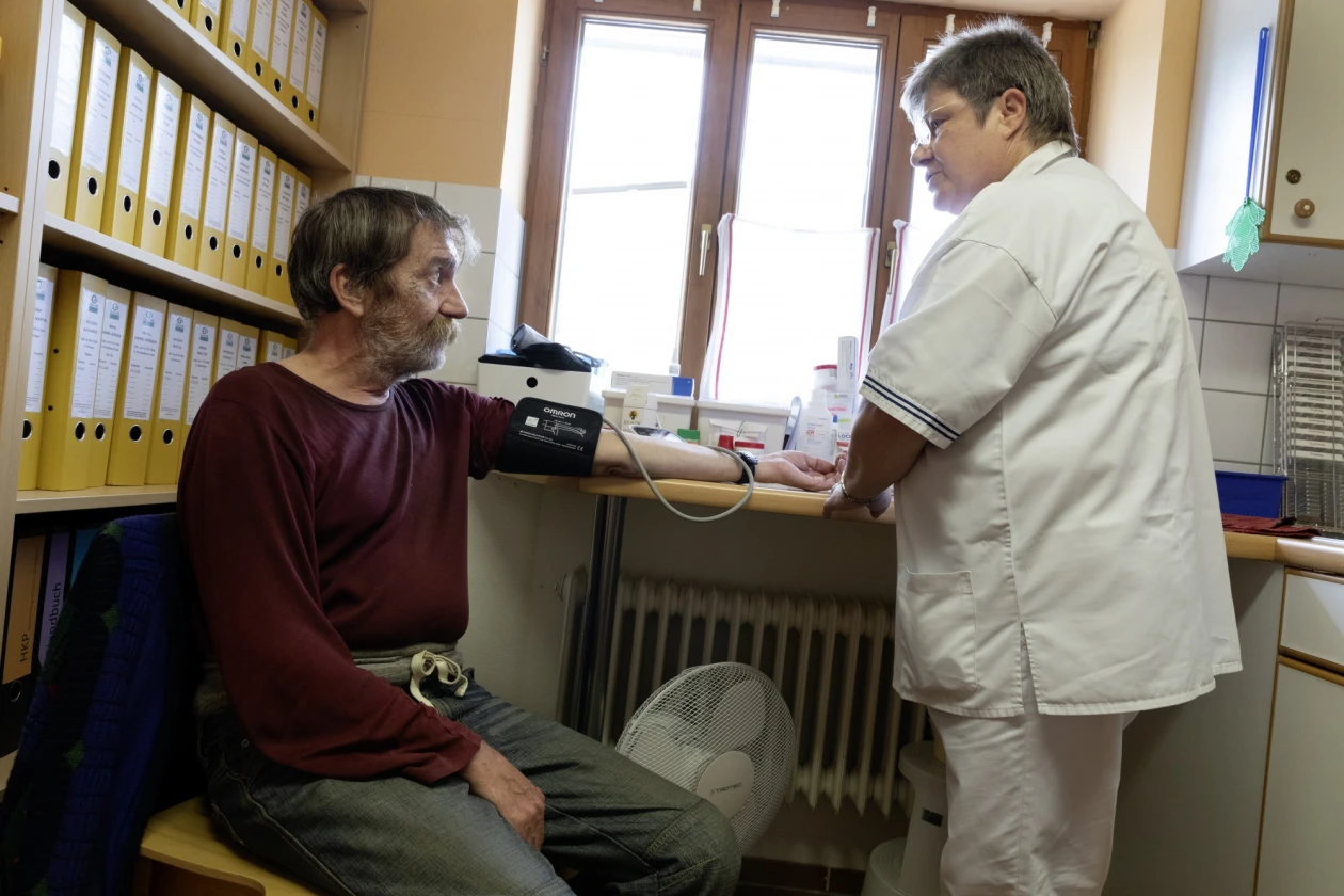 Ein Mann sitzt auf einem Stuhl in einem Raum, in dem ein Regal mit vielen Ordnern steht, w&auml;hrend eine Frau in wei&szlig;em Kittel eine Blutdruckmessung durchf&uuml;hrt.