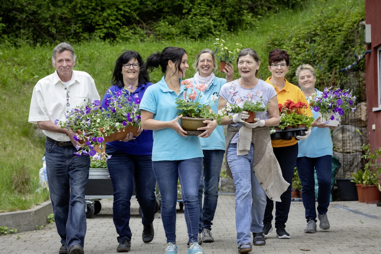 Sieben l&auml;chelnde Menschen tragen verschiedene Blument&ouml;pfe und -arrangements, w&auml;hrend sie zusammen auf einem Weg vor einem gr&uuml;nen Hintergrund gehen.