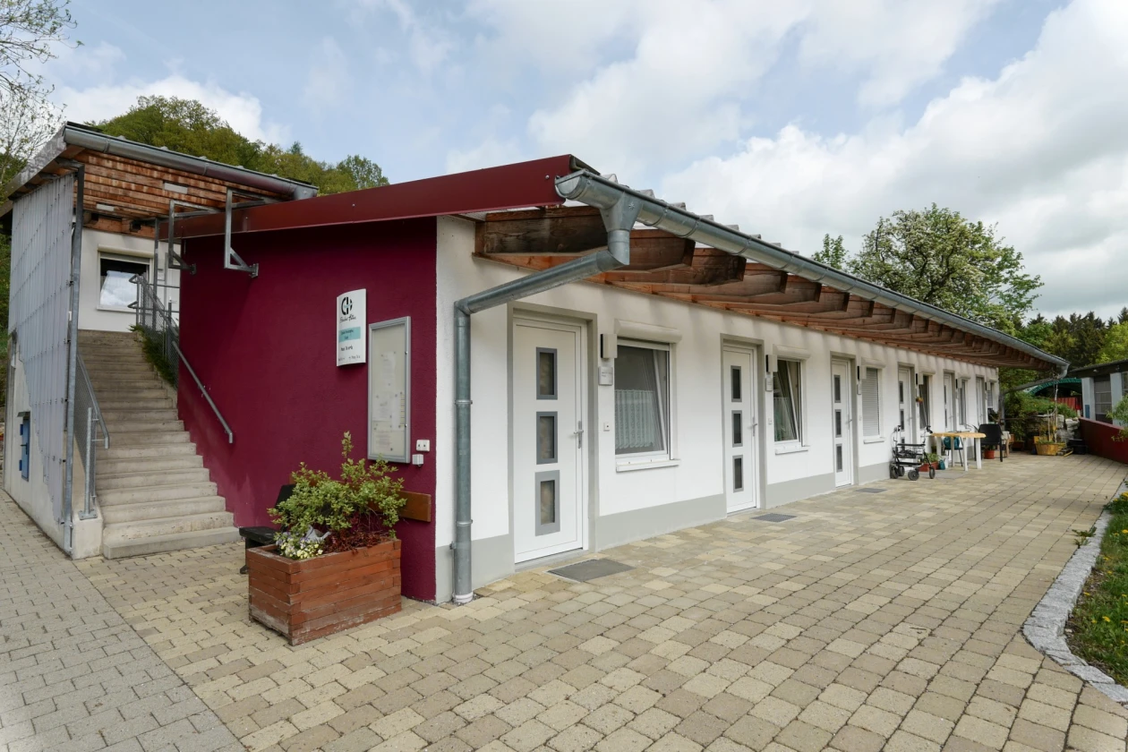 Modernes Geb&auml;ude in Bordeauxrot und Wei&szlig; mit Betonpflasterweg, Holz&uuml;berdachung und Treppe. Mehrere Fenster und eine Holzkiste mit Pflanzen stehen vor der Fassade. Ruhige Umgebung mit B&auml;umen und leicht bew&ouml;lktem Himmel im Hintergrund.