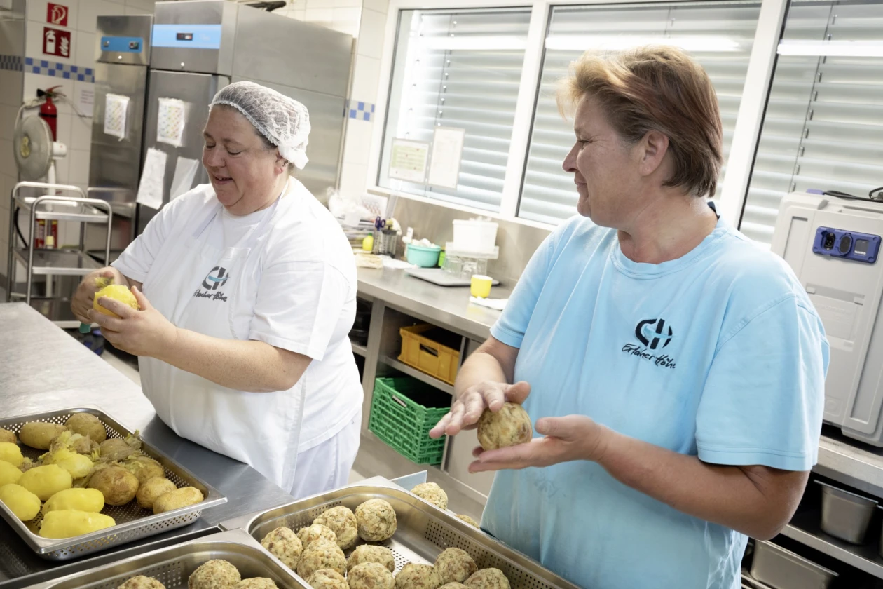 Zwei Frauen stehen in einer K&uuml;che. Eine Frau tr&auml;gt ein wei&szlig;es T-Shirt, die Frau ein hellblaues T-Shirt, beide mit Logo der Erlacher H&ouml;he. Die linke Frau sch&auml;lt Kartoffeln, die rechte Frau formt Semmelkn&ouml;del. Sie stehen an einem Arbeitstisch mit Metallbeh&auml;ltern.