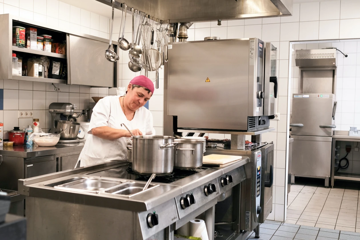 Eine Frau in wei&szlig;er Arbeitskleidung und rosa Haube steht an einem gro&szlig;en Edelstahl-K&uuml;chenblock mit T&ouml;pfen.