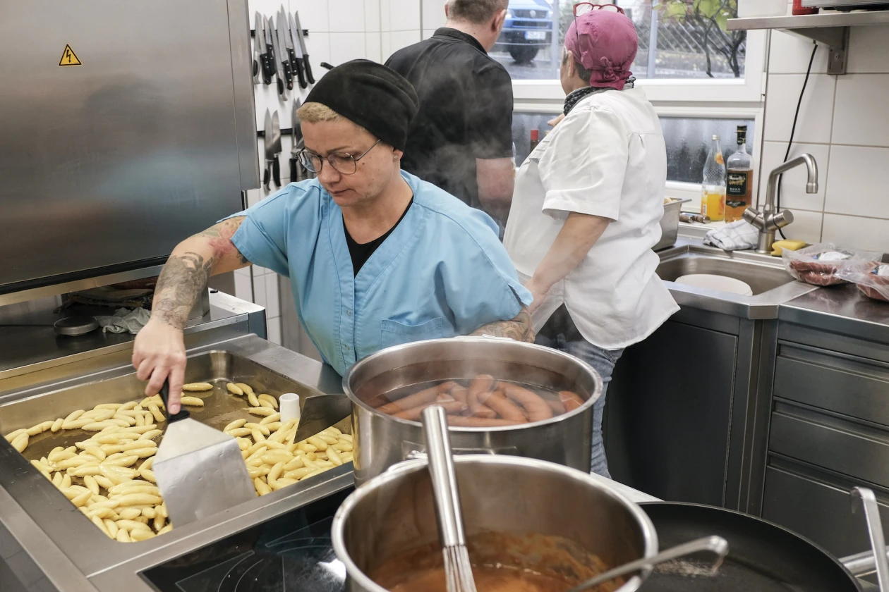 Drei Personen in einer Gro&szlig;k&uuml;che. Eine Frau in blauer Sch&uuml;rze, Haube und Brille br&auml;t Schupfnudel in einem gro&szlig;en Br&auml;ter. Im Vordergrund sind zwei gro&szlig;e Kocht&ouml;pfe mit verschiedenen Speisen auf dem Herd stehen. Eine weitere Frau mit wei&szlig;em Kassack steht im Hintergrund an der Sp&uuml;le, einem Mann in schwarzem Oberteil zugewandt.