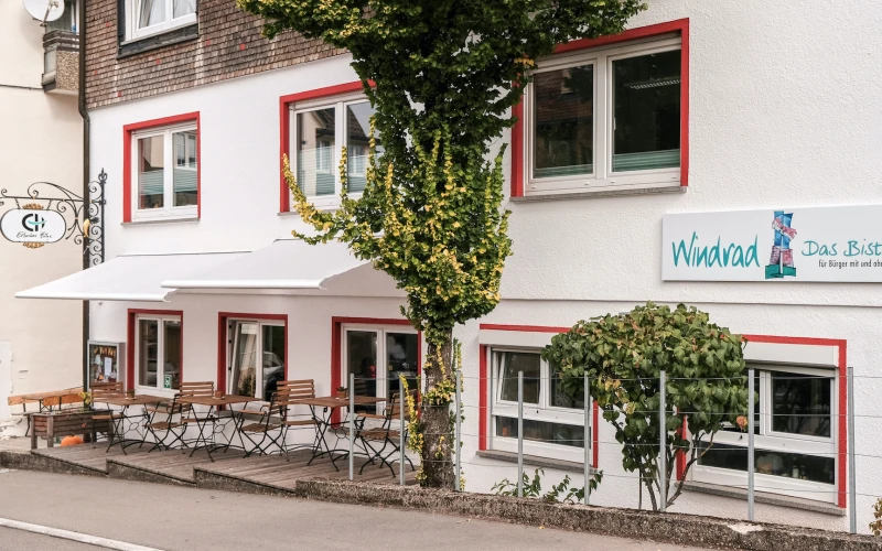 Wei&szlig;-rotes Geb&auml;ude mit Au&szlig;enbereich, bestehend aus Bistrotischen mit St&uuml;hlen, teilweise von einem Baum beschattet. An der Fassade h&auml;ngt ein Schild mit dem Schriftzug "Windrad" Das Bistro." Die Fenster haben rote Rahmen.