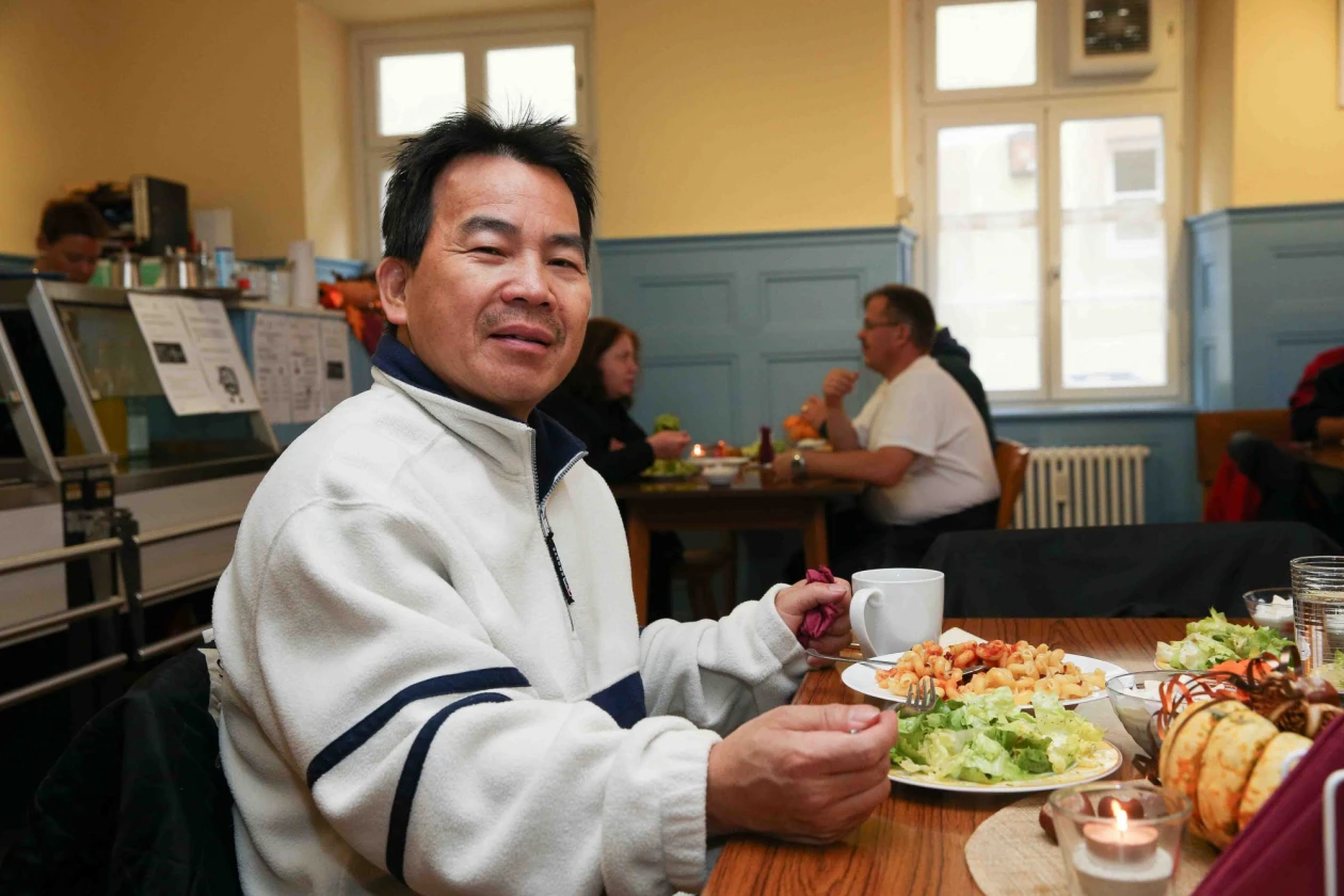 Ein asiatisch aussehender Mann in einem wei&szlig;en Pullover sitzt an einem Tisch mit Essen und einer Tasse. Er l&auml;chelt in die Kamera, w&auml;hrend im Hintergrund weitere Personen an Tischen sitzen.