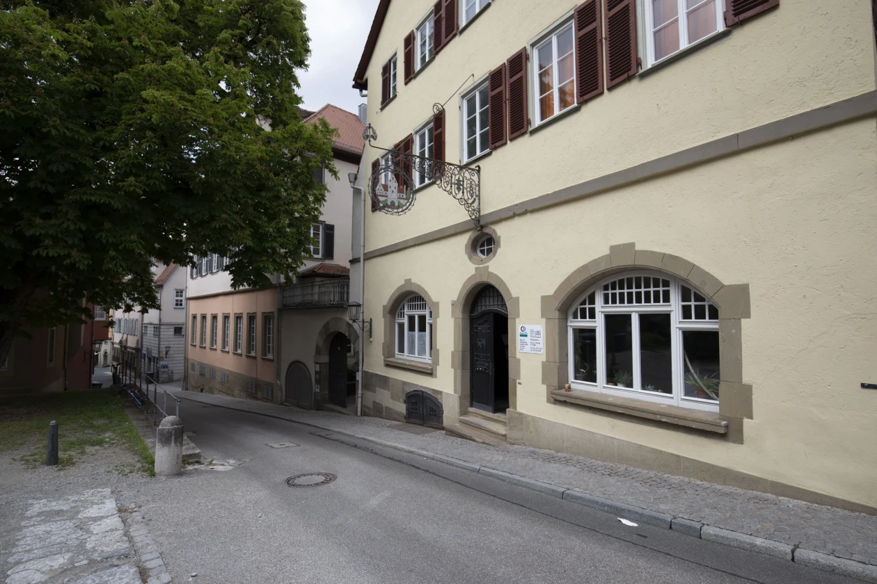 Schmale Altstadtstra&szlig;e mit historischen Geb&auml;uden in hellem Sandstein, ges&auml;umt von einem gro&szlig;en Baum links und klassizistischer Architektur mit dunkelbraunen Fensterl&auml;den und Verzierungen. An der Fassade h&auml;ngt ein Schild mit der Aufschrift "Zur Schuppachburg".