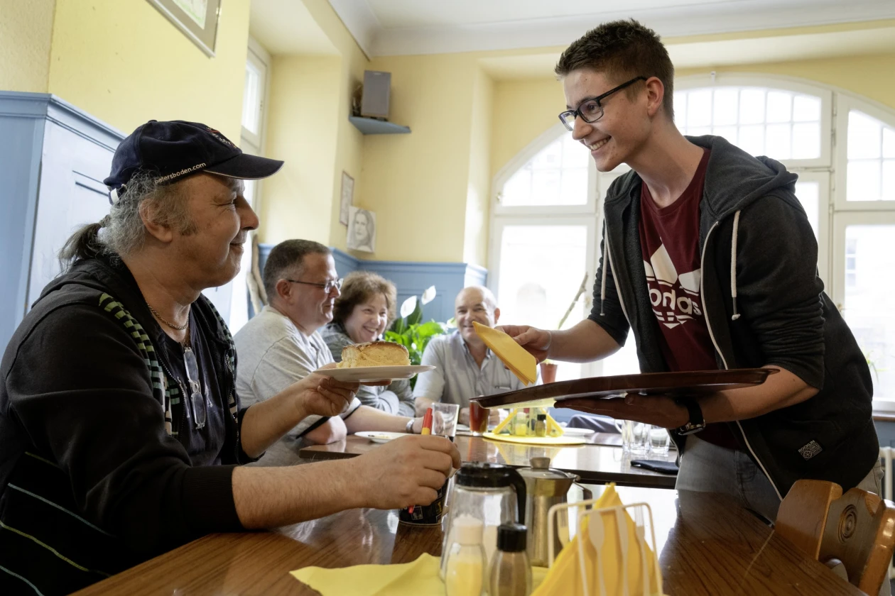 Ein junger Mann serviert einem Mann mit M&uuml;tze und Zopf Essen an einem Tisch.. Die Szene spielt in einem hellen, freundlichen Raum mit Holzm&ouml;beln und gro&szlig;en Fenstern. Im Hintergrund sind weitere G&auml;ste an einem Tisch zu sehen.