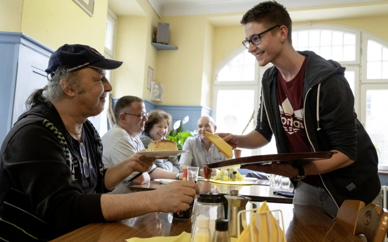 Ein junger Mann serviert einem Mann mit M&uuml;tze und Zopf Essen an einem Tisch.. Die Szene spielt in einem hellen, freundlichen Raum mit Holzm&ouml;beln und gro&szlig;en Fenstern. Im Hintergrund sind weitere G&auml;ste an einem Tisch zu sehen.