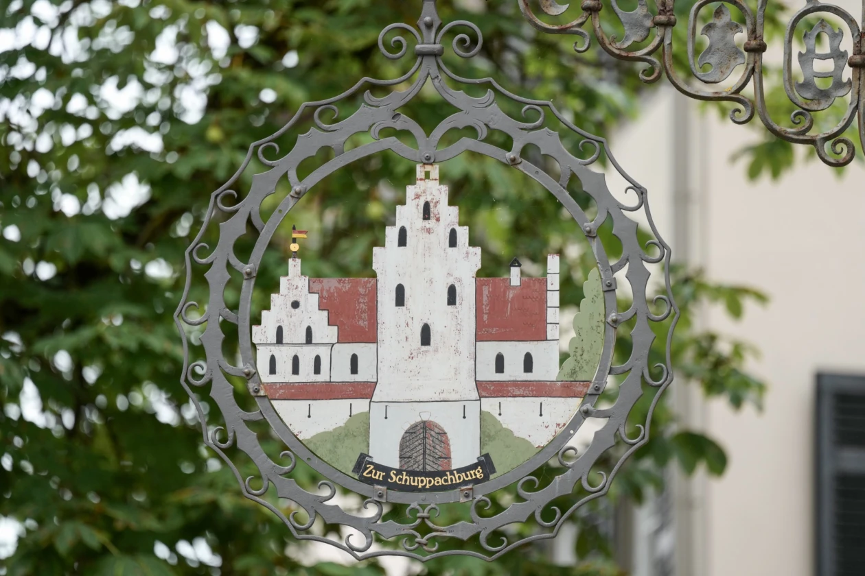 Schmiedeeisernes, rundes Ornament mit detaillierter Darstellung einer historischen Burg, gerahmt von geschwungenen Metallverzierungen vor einem unscharfen gr&uuml;nen Hintergrund mit Bl&auml;ttern. Auf dem Schild steht "Zur Schuppachburg".