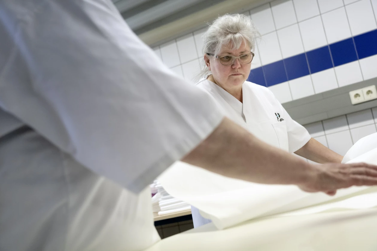 Eine Frau in wei&szlig;er Arbeitskleidung arbeitet sorgf&auml;ltig an einer Arbeitsfl&auml;che in einer W&auml;scherei. Sie legt mit einer weiteren Person, die  im Vordergrund zu sehen ist, ein W&auml;schest&uuml;ck zusammen.