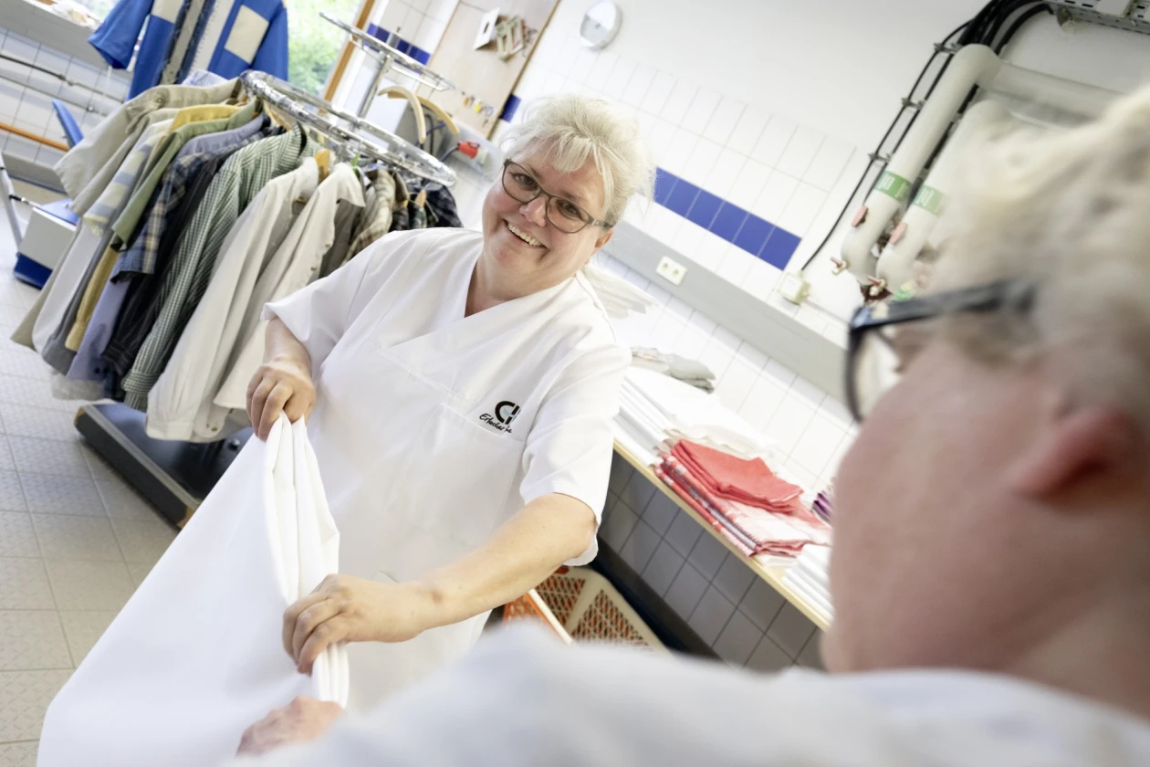 Eine Frau in wei&szlig;er Berufskleidung in einer W&auml;scherei l&auml;chelt in die Kamera. Sie legt zusammen mit einer weiteren Person im Vordergrund ein wei&szlig;es Textilst&uuml;ck zusammen. Im Hintergrund h&auml;ngen Kleidungst&uuml;cke auf einem Kleiderst&auml;nder.