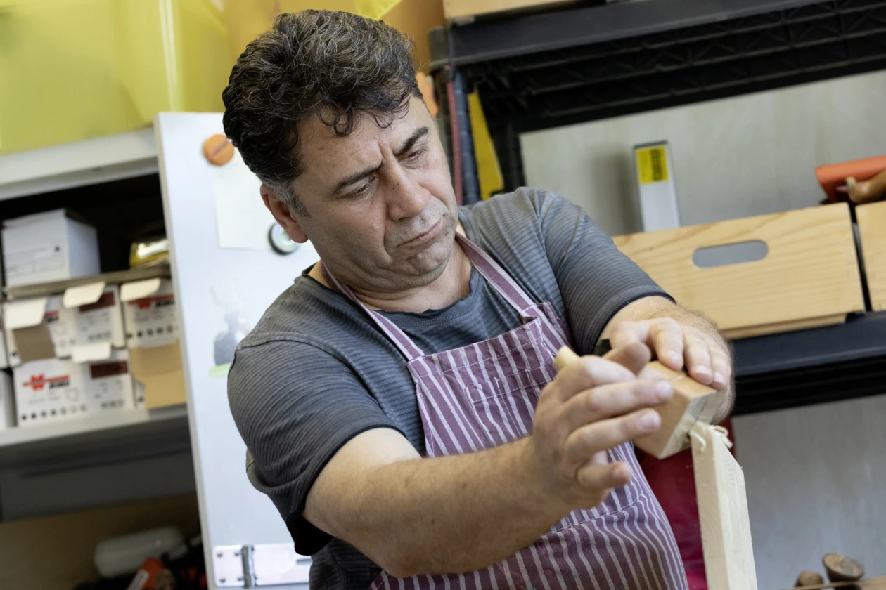 Ein Mann in gestreifter Sch&uuml;rze bei der Arbeit an einem Holzst&uuml;ck. Im Hintergrund sind weitere Materialien zu sehen.