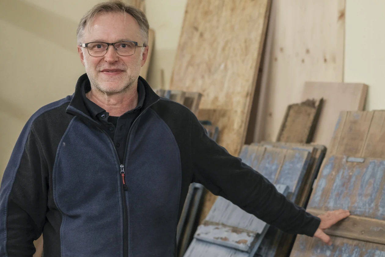 Ein Mann mit Brille und dunkler Jacke blickt in die Kamera. Er steht vor Holzbrettern, die an die Wand gelehnt sind.
