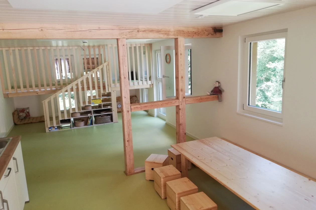 Ein Raum mit zwei Fenster, gr&uuml;nem Boden, Holzbalken und einer Treppe mit Gel&auml;nder. Holzw&uuml;rfel dienen als Sitzgelegenheit an einem gro&szlig;en Holztisch.