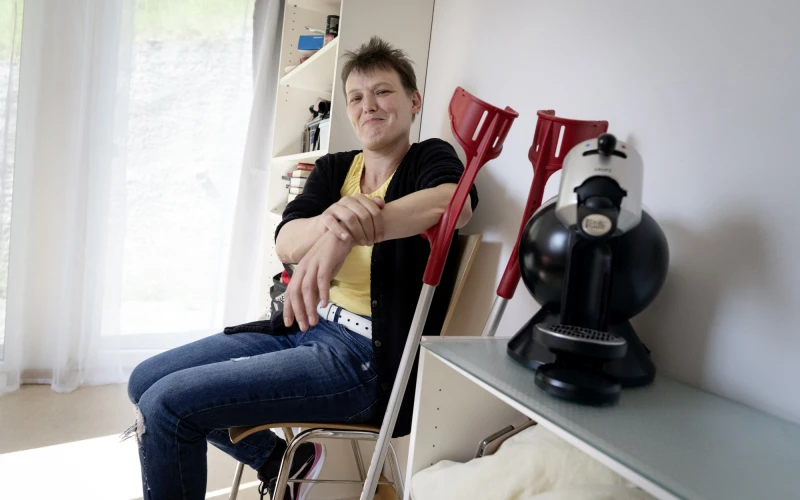 Eine Frau sitzt l&auml;chelnd auf einem Stuhl, neben sich zwei rote Kr&uuml;cken, links von ihr ein hohes wei&szlig;es Regal, rechts von ihr steht eine Kaffeemaschine auf einem kleinen Regal.