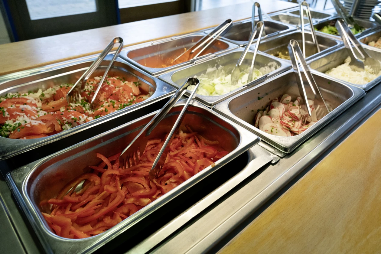 Blick auf ein Salatbuffet mit verschiedene Metallbeh&auml;lter mit verschiedenen Salaten und Paprikastreifen. Jeder Beh&auml;lter ist mit einer Salatzange best&uuml;ckt.