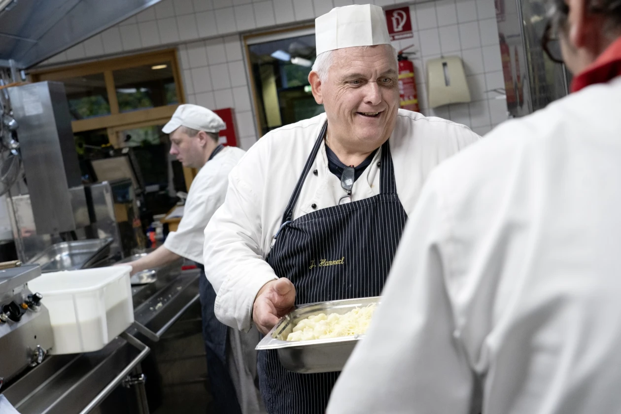 &Auml;lterer Koch in wei&szlig;er Kochjacke und gestreifter Sch&uuml;rze in einer K&uuml;che. L&auml;chelnd h&auml;lt er eine gro&szlig;e Auflaufform. Im Hintergrund sind weitere K&ouml;che bei der Arbeit zu sehen.