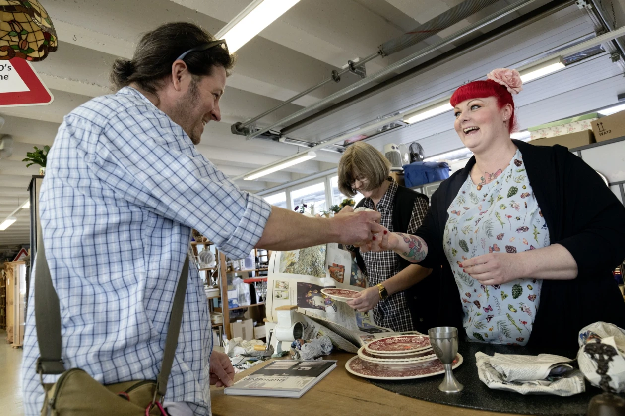 Es sind insgesamt 3 Personen zu sehen. Der Mann in einem karierten Hemd reicht einer l&auml;chelnden Frau mit roten Haaren einen Gegenstand &uuml;ber einen Tresen in einem Arbeitsbereich. Die andere Frau nebendran hat einen Teller in der Hand und schaut runter.