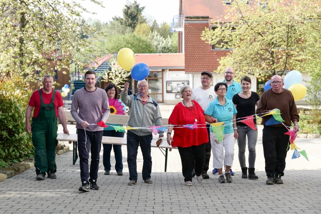 Gruppe von Menschen unterschiedlichen Alters und Geschlechts mit Luftballons und Wimpeln auf einem Pflasterweg, die gemeinsam an einer Veranstaltung oder einem Fest teilzunehmen scheinen, vor einem Backsteingeb&auml;ude und B&auml;umen.