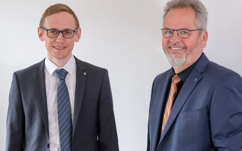 Zwei M&auml;nner in dunklen Anz&uuml;gen, Krawatten und Brillen, stehen vor einem hellen Hintergrund.