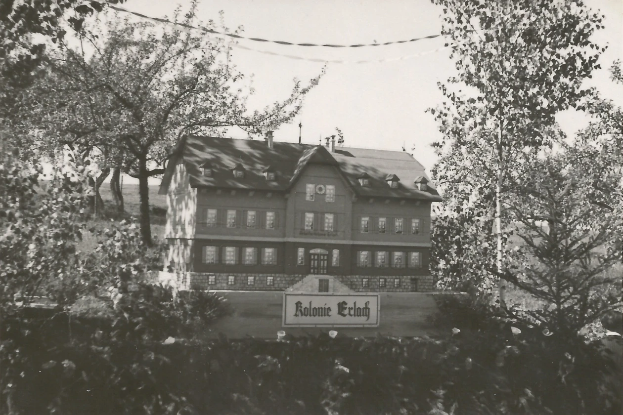 Historisches Schwarzwei&szlig;foto eines gro&szlig;en mehrgeschossigen Geb&auml;udes, umgeben von B&auml;umen, mit dem Schriftzug "Kolonie Erlach" im Vordergrund.
