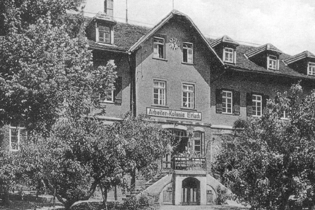 Historisches Schwarzwei&szlig;foto eines gro&szlig;en Backsteinhauses mit mehreren Stockwerken, umgeben von B&auml;umen und Str&auml;uchern, mit einer Treppe zum Haupteingang und einer beschrifteten Fassade "Arbeiter-Kolonie Erlach".