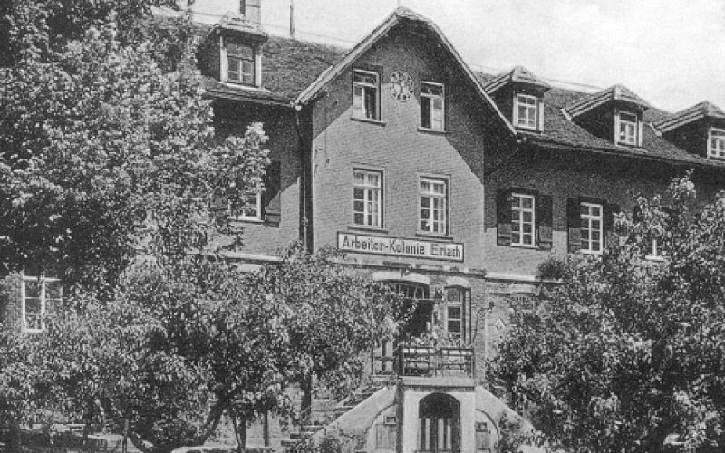 Historisches Schwarzwei&szlig;foto eines gro&szlig;en Backsteinhauses mit mehreren Stockwerken, umgeben von B&auml;umen und Str&auml;uchern, mit einer Treppe zum Haupteingang und einer beschrifteten Fassade "Arbeiter-Kolonie Erlach".
