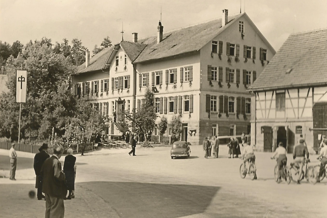 Historische Schwarzwei&szlig;aufnahme eines Dorfplatzes mit mehreren Fachwerkh&auml;usern, B&auml;umen und Personen, die zu Fu&szlig; oder mit Fahrr&auml;dern unterwegs sind, vor einem Geb&auml;ude mit einer Fahne.
