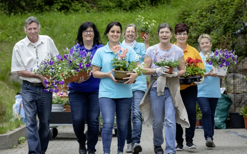Sieben Personen gehen in einer Reihe, jeder tr&auml;gt einen Blumentopf und l&auml;chelt dabei. Im Hintergrund ist eine gr&uuml;ne Gartenumgebung zu sehen.