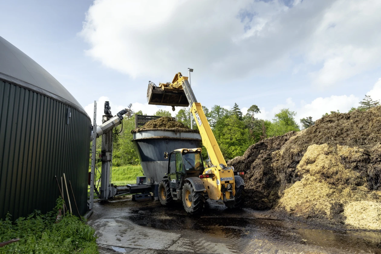 Landwirtschaftlicher Teleskoplader l&auml;dt Biomasse in eine gro&szlig;e Mulde neben einer Biogasanlage. Die Maschine steht auf einer feuchten Zufahrt mit einem gro&szlig;en Misthaufen. Im Hintergrund ist eine gr&uuml;ne Wiese zu sehen.