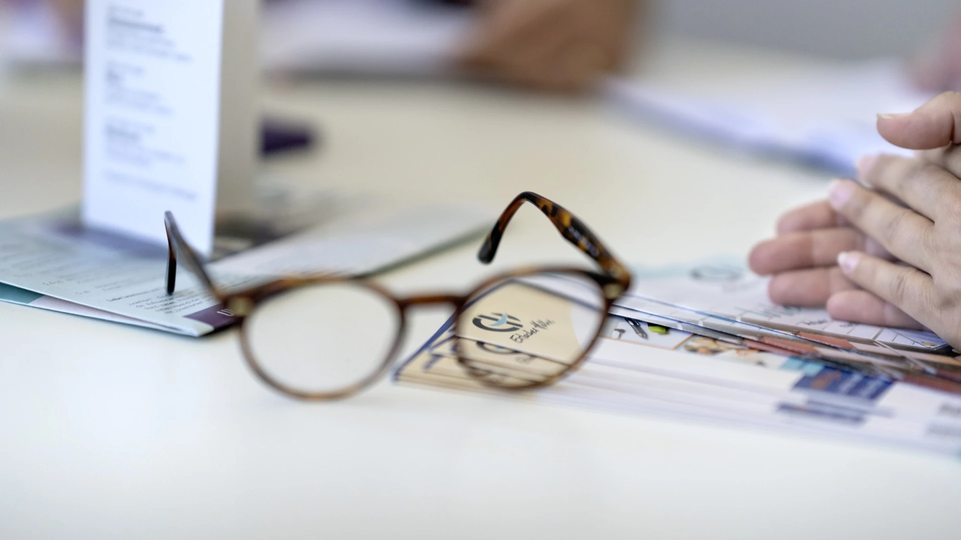 Braune Lesebrille auf einem Papierdokument mit EH Logo und unscharfem Hintergrund, auf dem eine Hand teilweise zu sehen ist. Die Brille liegt leicht schr&auml;g und reflektiert Licht auf einem Arbeitstisch.